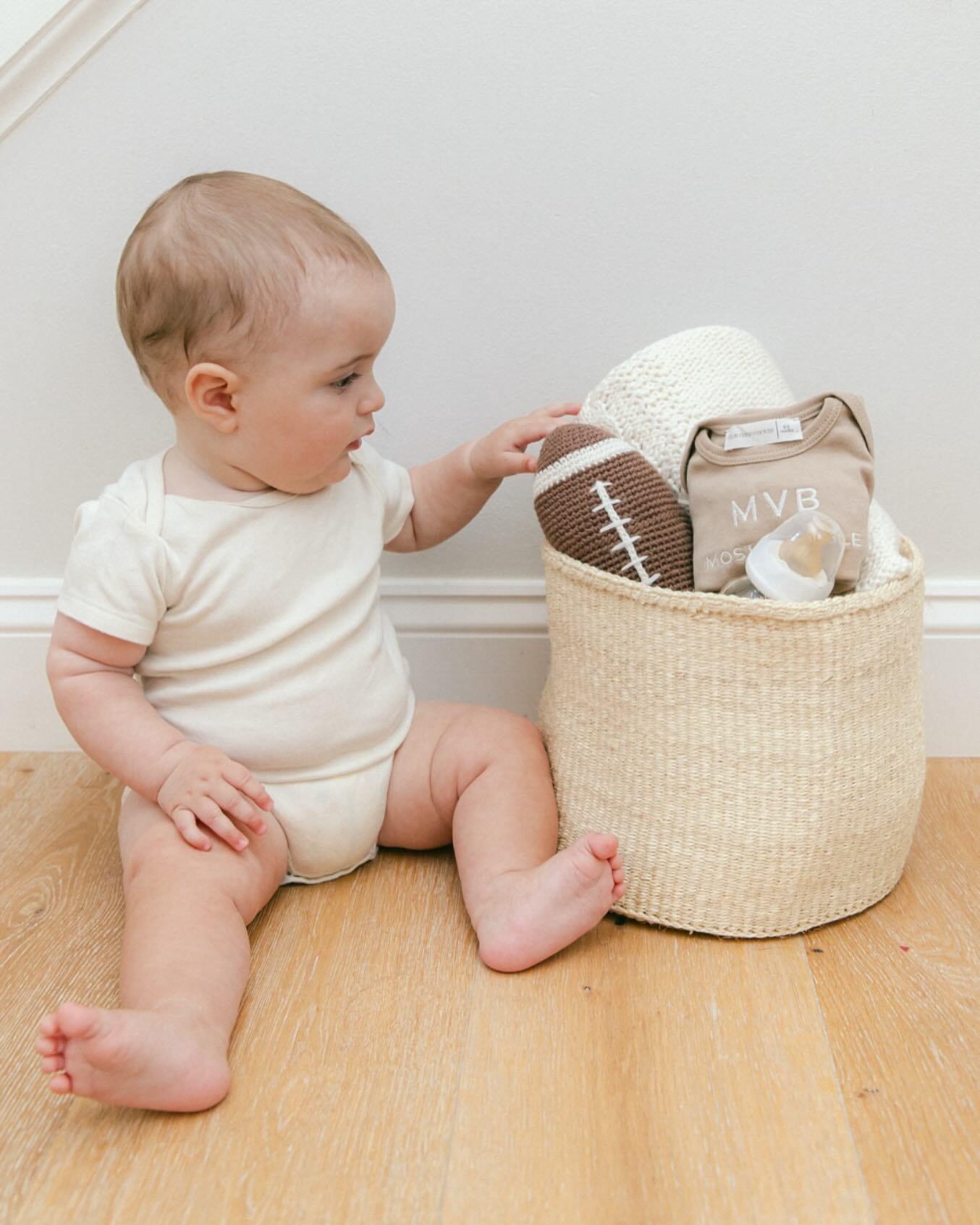  Baby sitting on wooden floor reaching into Our Green House basket filled with a football toy, blanket, and baby outfit.