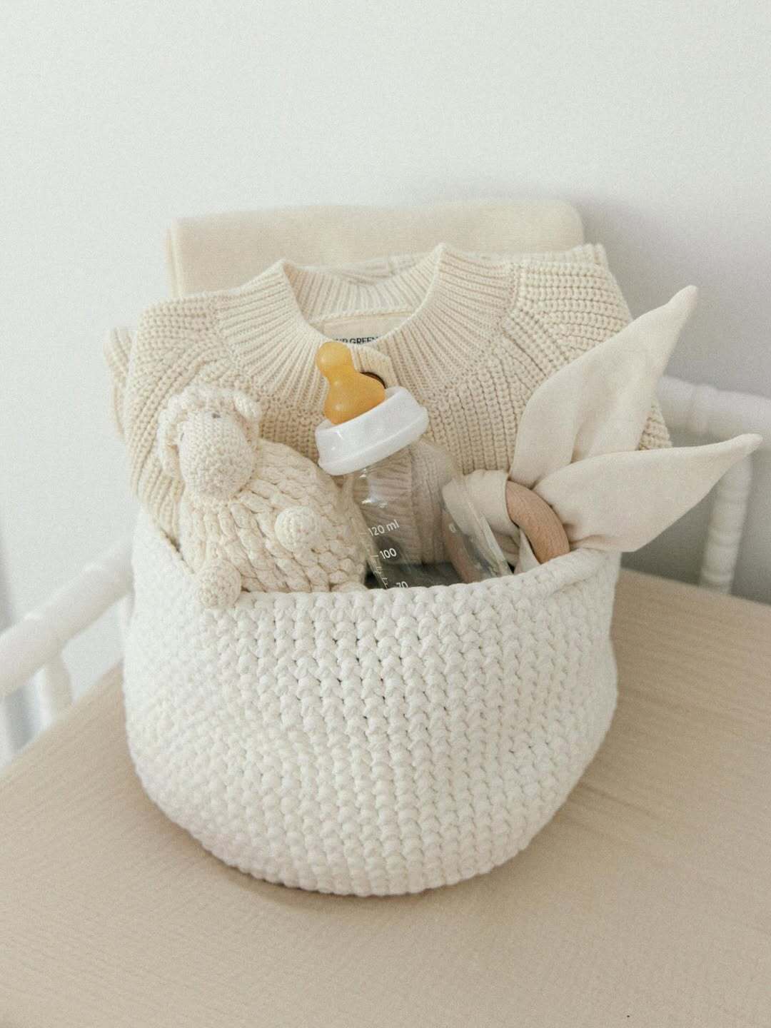 Crocheted basket filled with baby sweater, plush toy, bottle, and wooden teether from Our Green House.