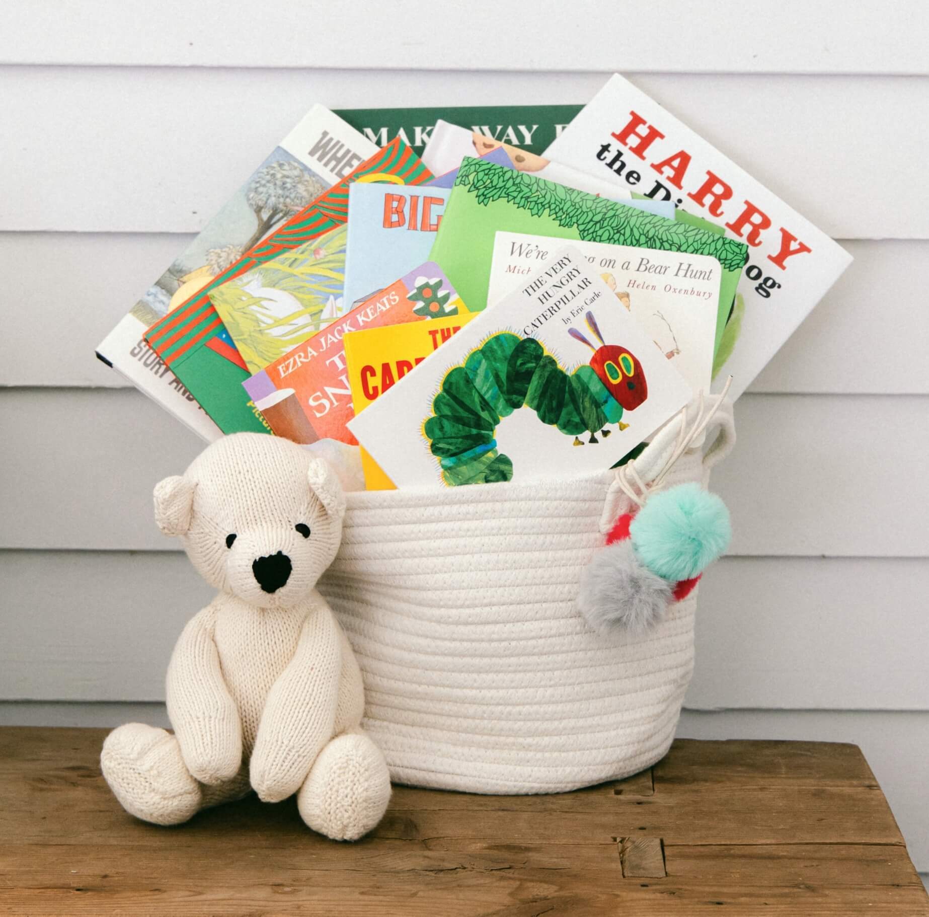 White cotton rope basket filled with children's books and a knitted white bear in front.