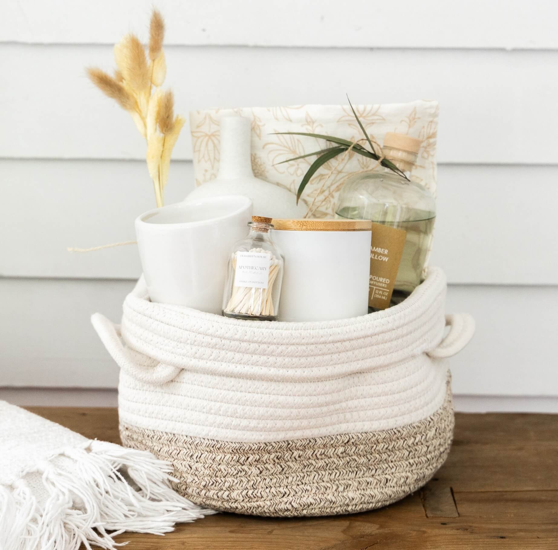 White rope basket holding dried flowers, a tea tin, candle, small jar, card, and stacked blankets as a self-care gift set.