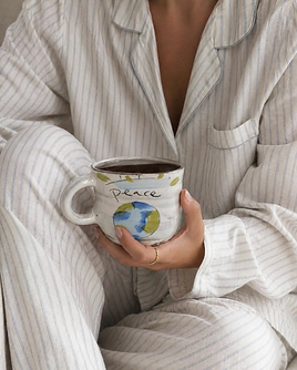 Person in striped linen pajamas holding a hand-painted “Peace” ceramic mug from Our Green House.