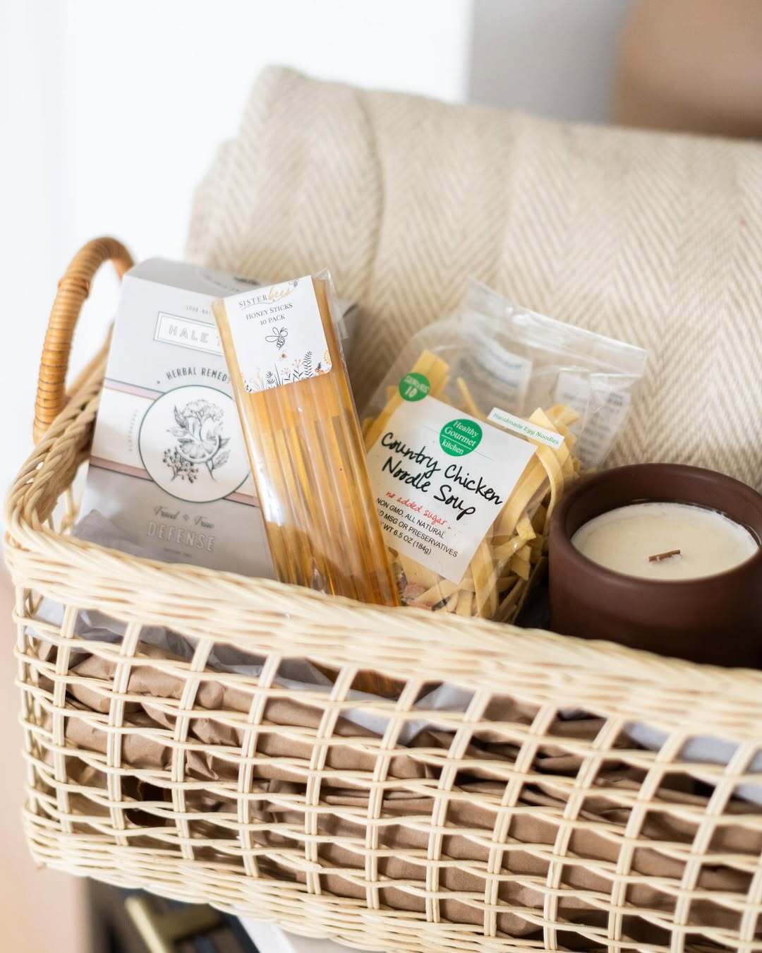 Wicker gift basket from Our Greenhouse with pasta, herbal tea, and a candle on a cozy blanket.