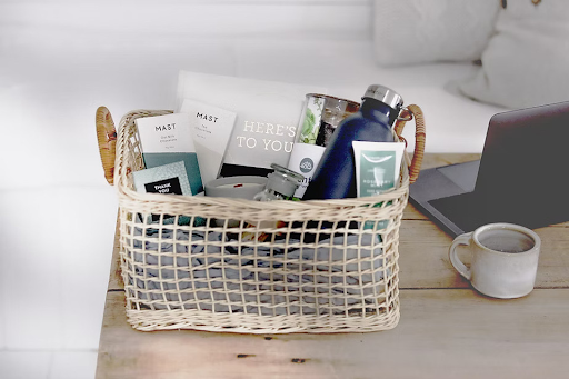 Woven basket care package on a desk with a laptop and mug, containing cards, a water bottle, and other items.